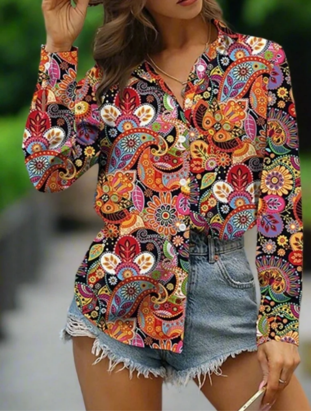 A woman wearing a vibrant, colourful floral-patterned blouse with long sleeves and a V-neck paired with denim shorts.