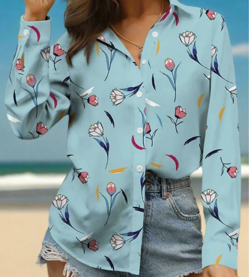 A woman in a light blue blouse featuring a delicate floral pattern with pink and white flowers, styled with rolled-up sleeves and denim shorts.