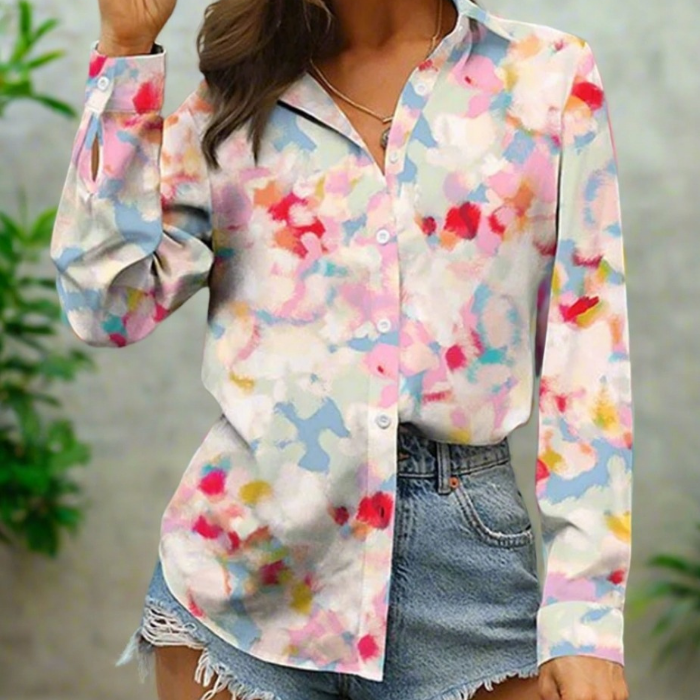 A woman showcasing a white blouse with a colourful abstract floral print, featuring long sleeves and a button-down front, paired with casual denim shorts.