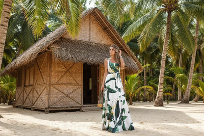 Woman in a leaf-patterned dress standing in front of a thatched-roof hut with palm trees in the background