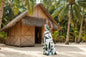 Woman in a leaf-patterned dress standing in front of a thatched-roof hut with palm trees in the background