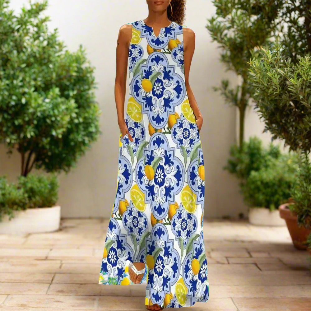 White sleeveless maxi dress with intricate blue floral and vine patterns, modelled indoors beside a potted plant