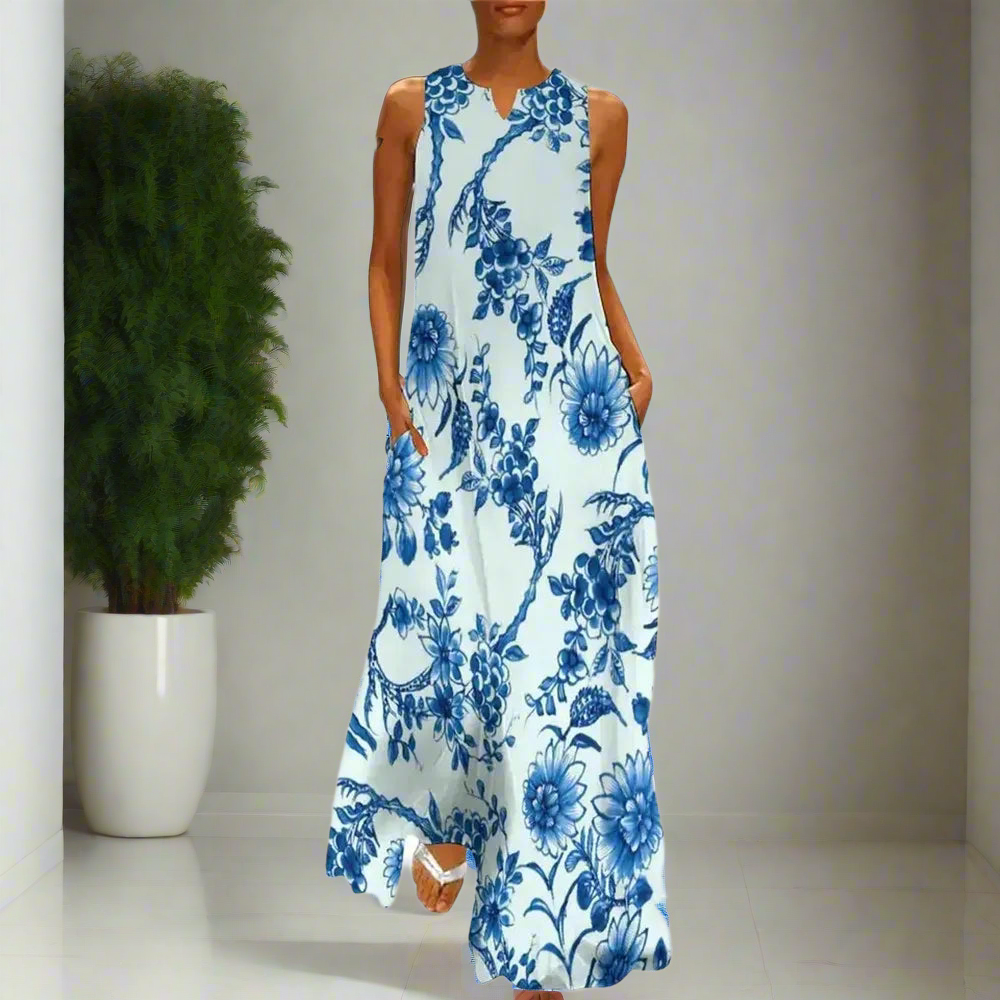 Elegant white maxi dress with blue floral print, sleeveless design, modelled indoors beside a tall potted plant