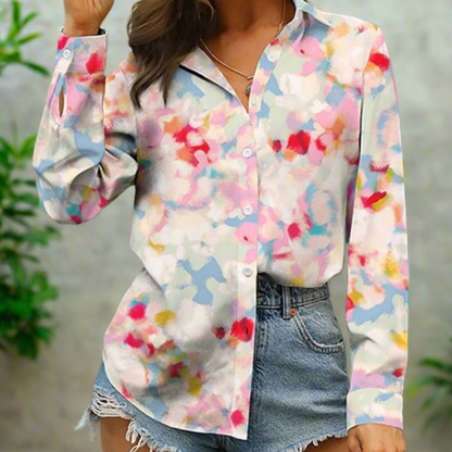 A woman showcasing a white blouse with a colourful abstract floral print, featuring long sleeves and a button-down front, paired with casual denim shorts.
