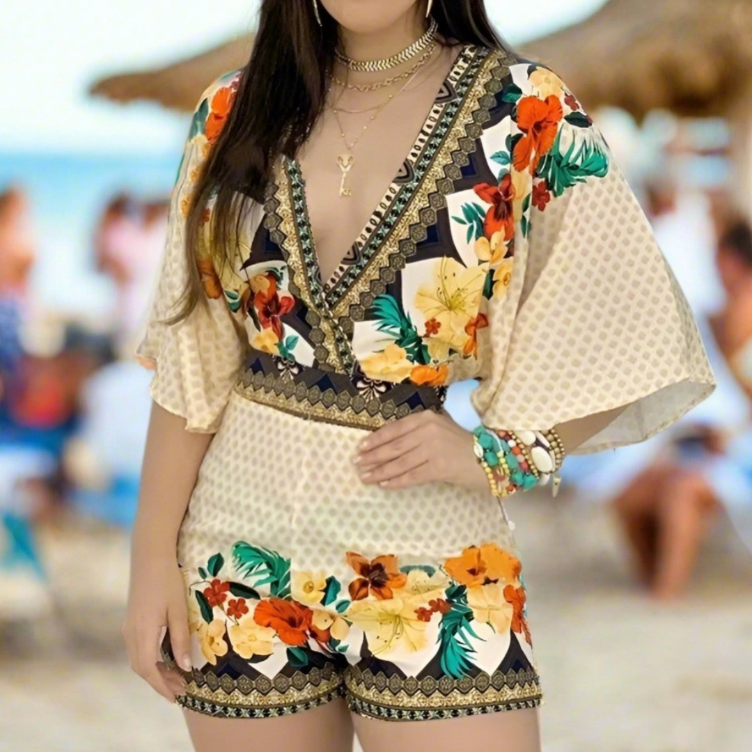 Woman wearing a cream floral romper with ruffle sleeves and shorts, standing near a beach hut.