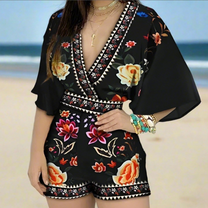 Woman wearing a black V-neck romper with multicolor floral patterns and wide sleeves, standing in an outdoor cafe setting