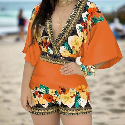 Woman in an orange V-neck romper with floral accents and wide sleeves, posing on a sandy beach