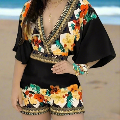 Woman wearing a black V-neck romper with gold-trimmed floral designs and wide sleeves, standing in a tropical beach setting