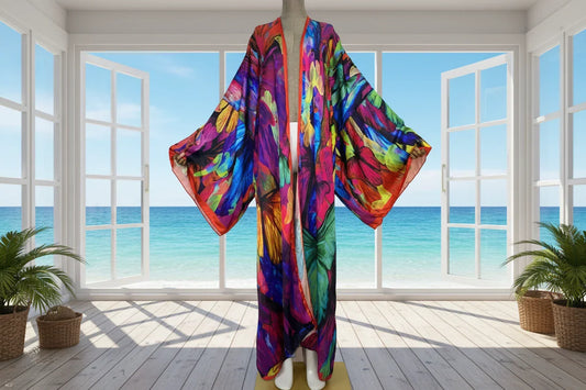 Woman wearing vibrant graffiti print long kimono gown standing on a sunlit balcony overlooking the ocean.