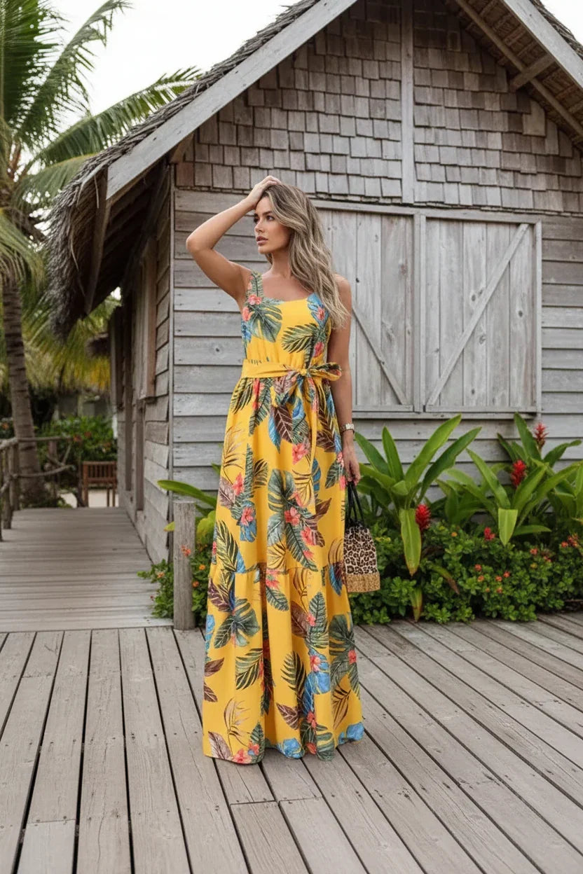 Woman in a yellow floral dress standing on a wooden deck with a rustic building and plants in the background.